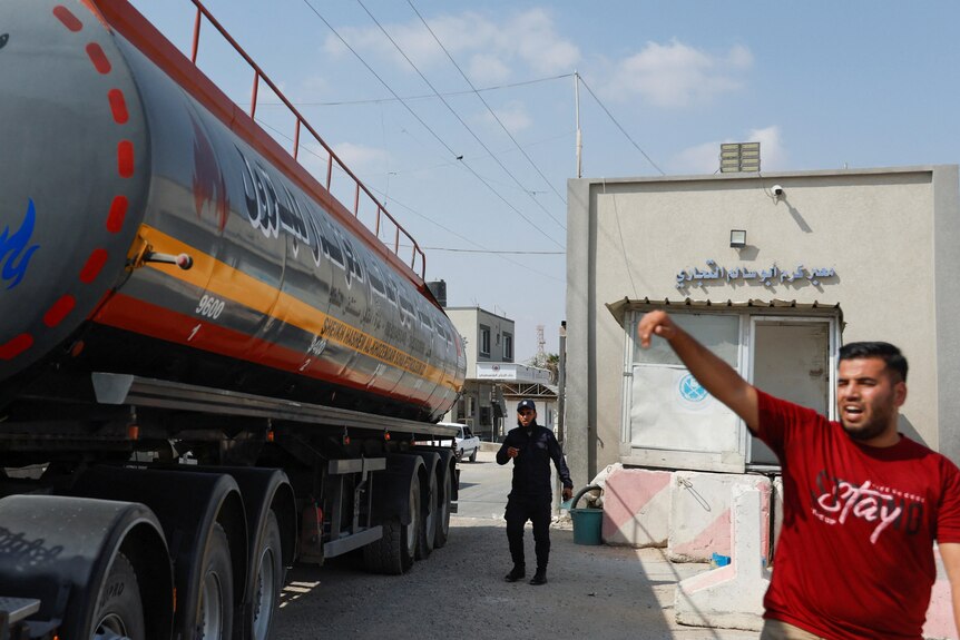 Man in red t-shirt holds arm up in gesture to motion to a fuel truck beside him.