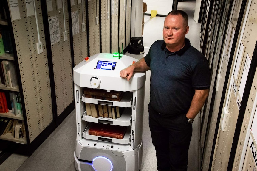 Stack attendant Paul Norden is standing next to an Automated Guided Vehicle among library stacks.