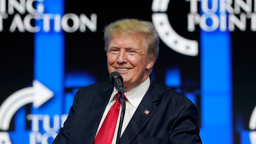 Donald Trump smiles while standing at a lectern with a microphone.