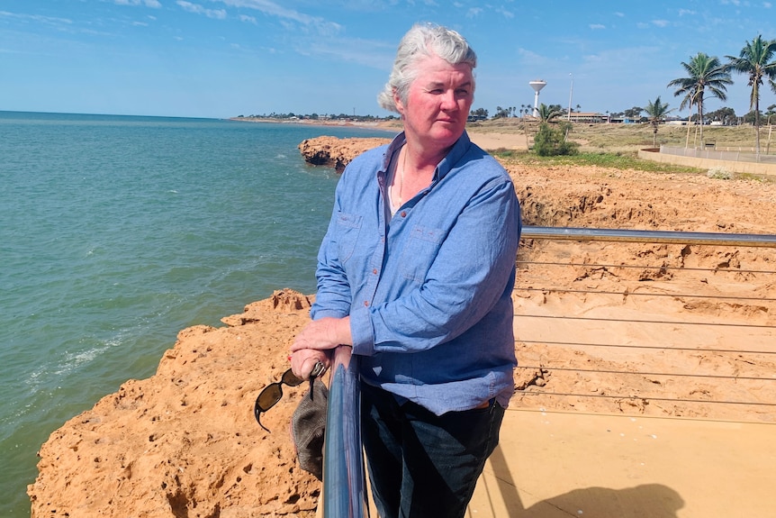 A grey-haired woman stands against a railing near the sea on a bright day.