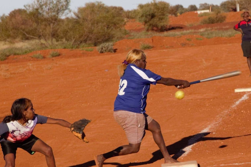 Women playing softball in red dirt.