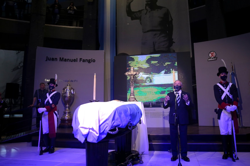 A mask-wearing man in a suit gestures as he speaks next to a flag-draped coffin flanked by two honour guards.