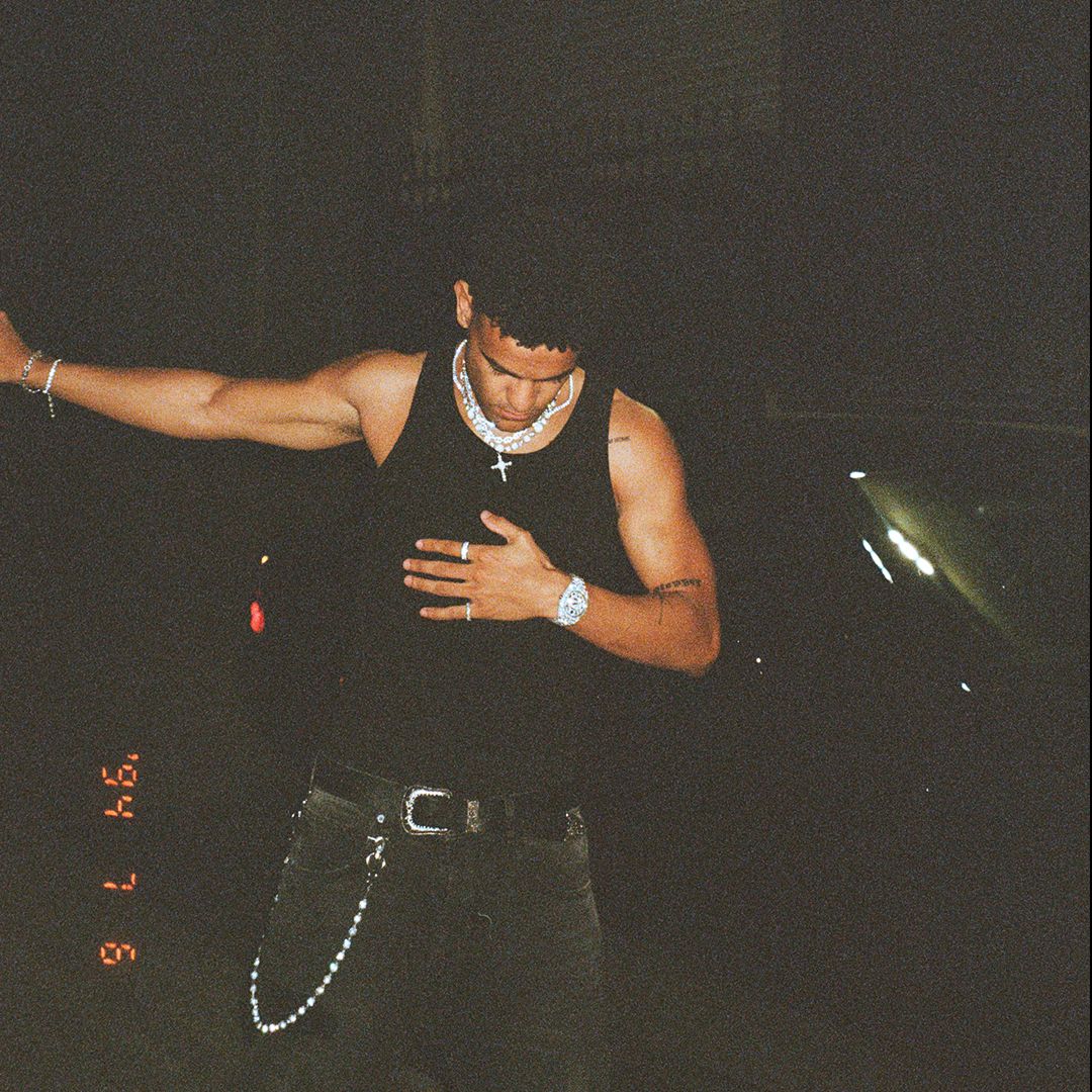 Faded film grade image of a man in a black singlet and pants with silver jewellery at night standing next to a black car