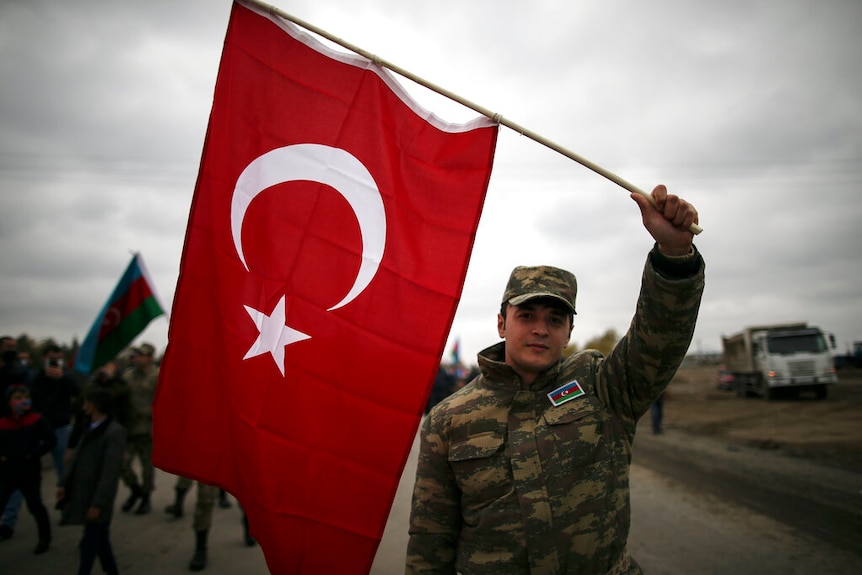 You view an Azerbaijani soldier holding the Turkish national flag amid a crowd of soldiers on an overcast day.