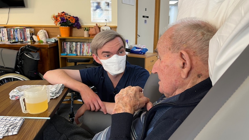 Ethan wearing scrubs and a face mask crouches down and speaks to aged care resident Bill Clark.