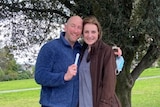 A man and a woman smile in a park holding a pregnancy test