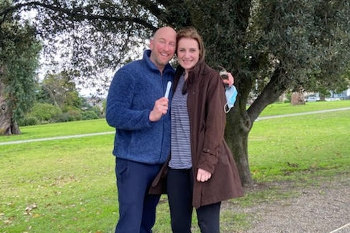 A man and a woman smile in a park holding a pregnancy test