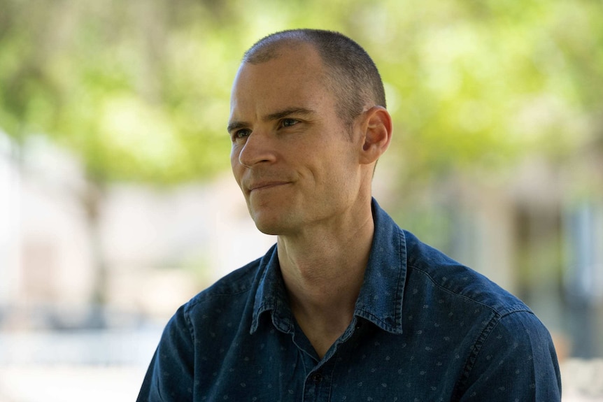 A man in a blue shirt in front of green trees.