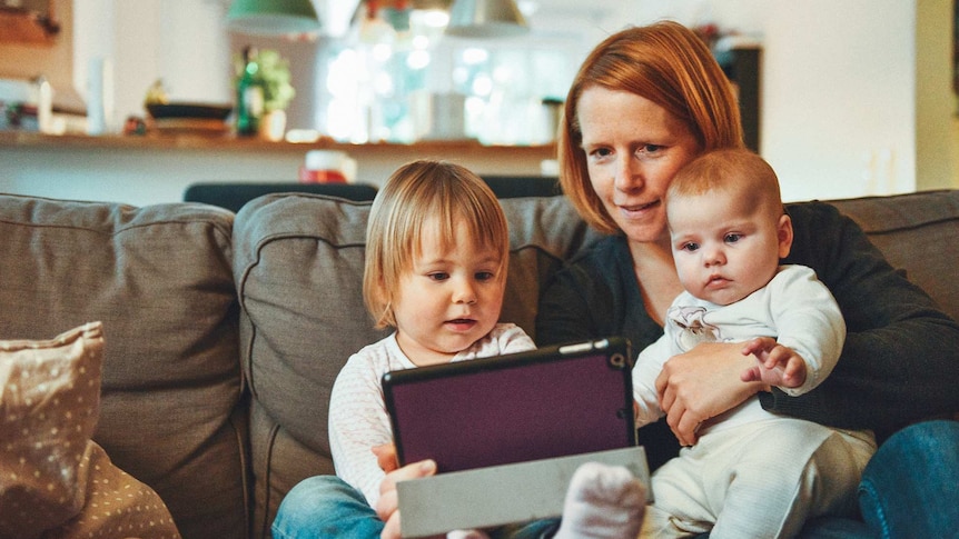 Woman holding a baby seated next to a toddler on the couch, all looking at a tablet device.