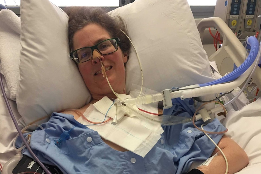A woman lies smiling in a hospital bed surrounded by tubes and machines.