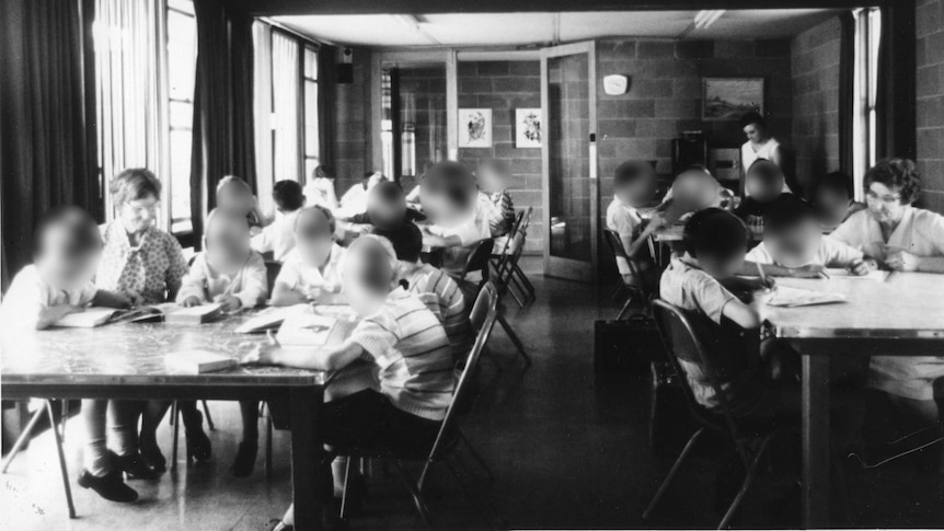 A black and white photo of boys and some adults sitting at tables doing school work.
