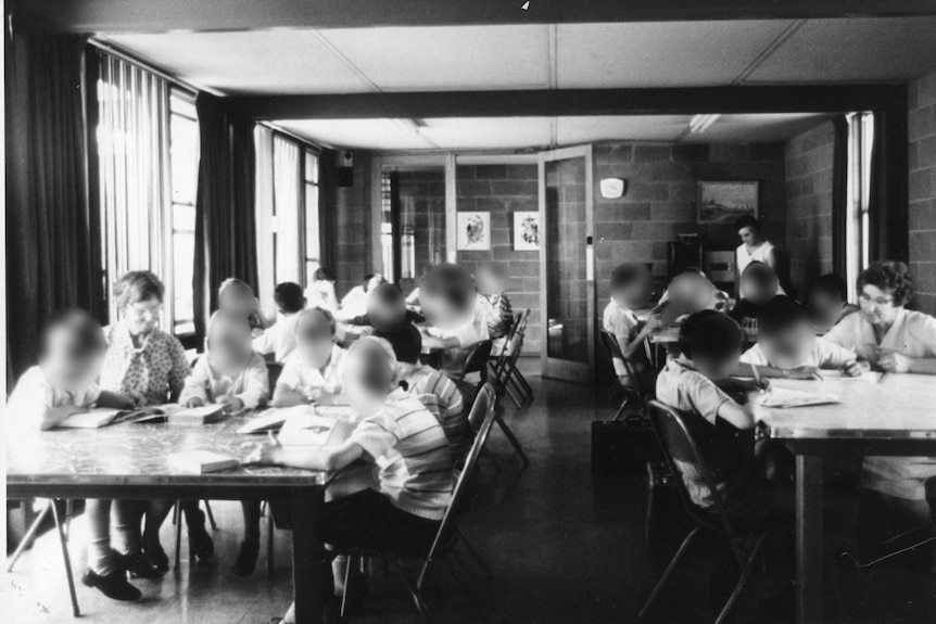 A black and white photo of boys and some adults sitting at tables doing school work.