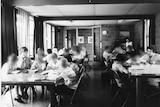 A black and white photo of boys and some adults sitting at tables doing school work.