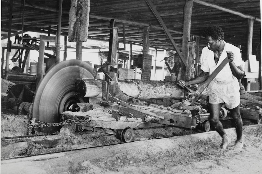 A monochrome showing a barefoot man manually pushing a log through a large cutting wheel.