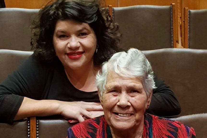 An elderly Indigenous Australian woman sits and smiles, with a younger woman sitting and smiling behind her