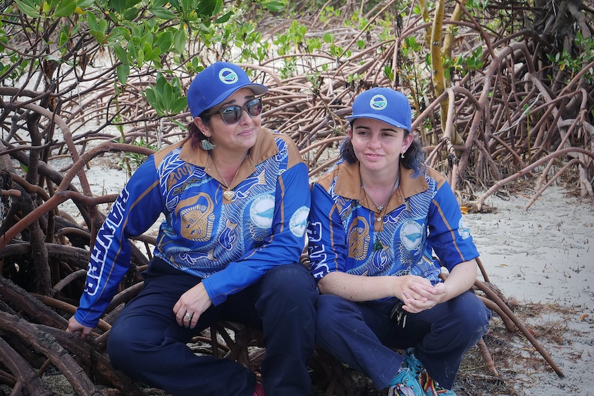 Two women in blue collared shirts and caps sit on some tree roots on a sandy beach