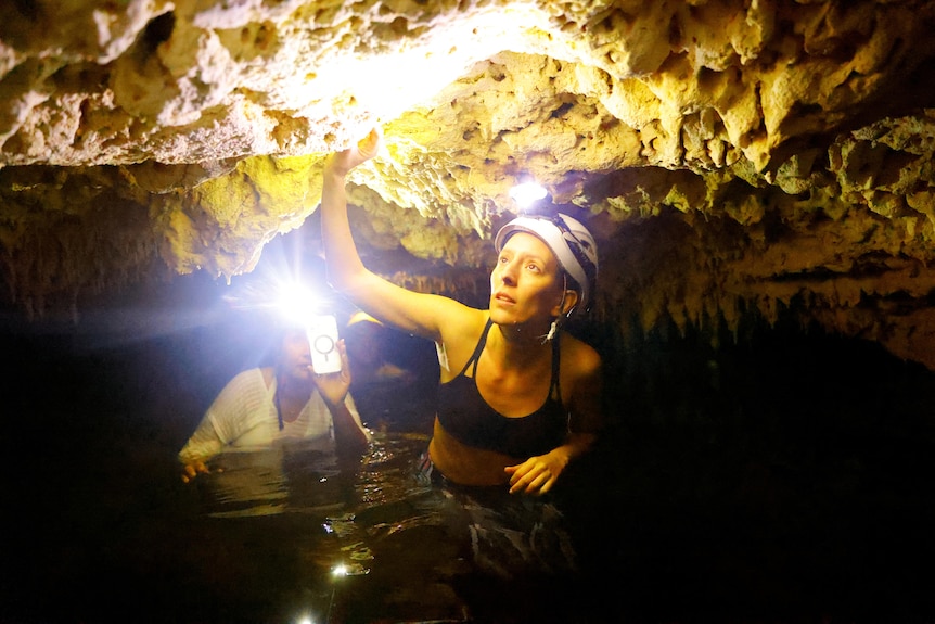 Two people in helmets walk through waist deep water inside a cave. 
