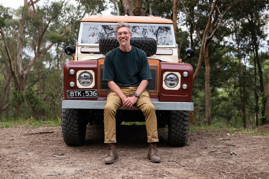A man sits on the front bumper of a vintage Land Rover, in a bush clearing