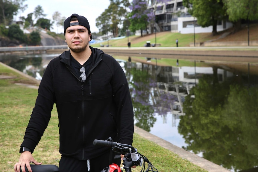 A man wearing a backwards cap and a hoody stands before a river holding a bicycle.