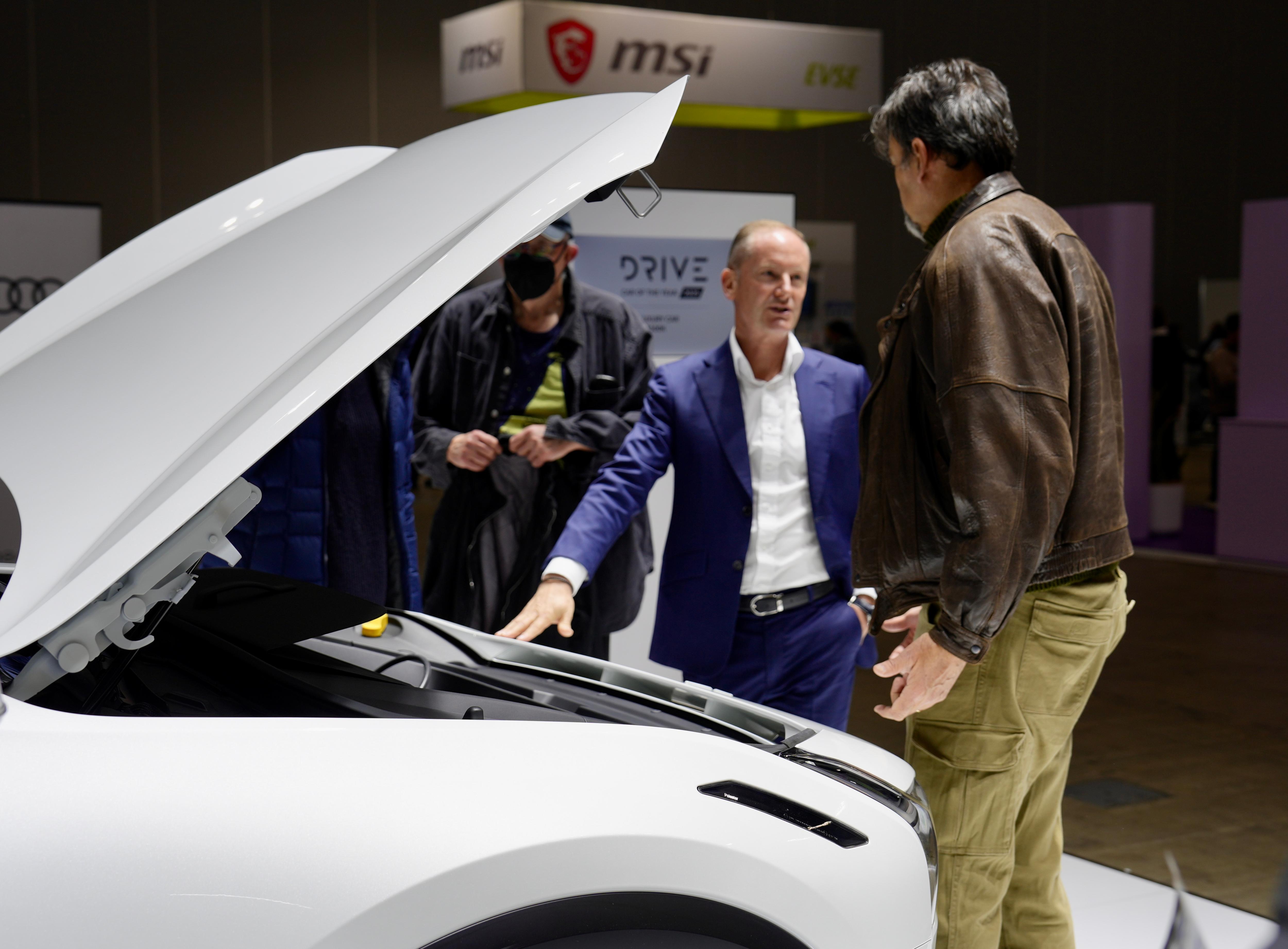 Two men talk next to a white car with its bonnet raised.