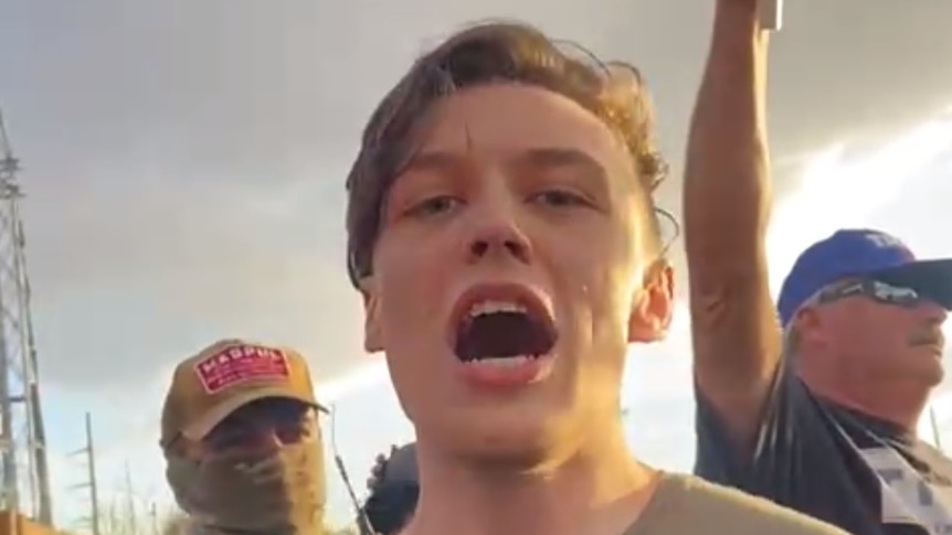 a young man with his mouth open stares at the camera with protesters behind him 