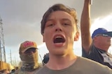 a young man with his mouth open stares at the camera with protesters behind him 
