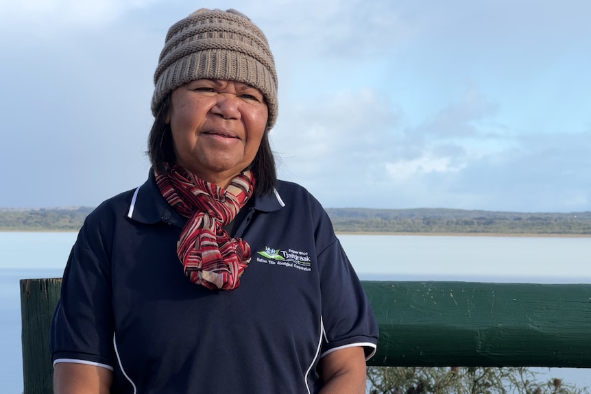 An older Indigenous woman wears a beanie, scarf, blue t-shirt with white logo. Lake behind her is light blue, smiles slightly.
