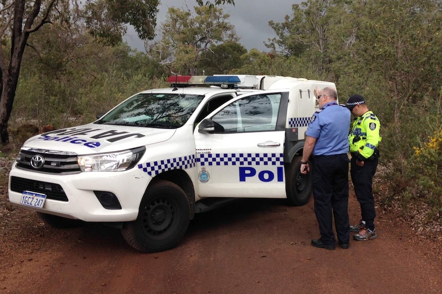 Two police officers stand next to a police vehicle on a dirt road in bushland