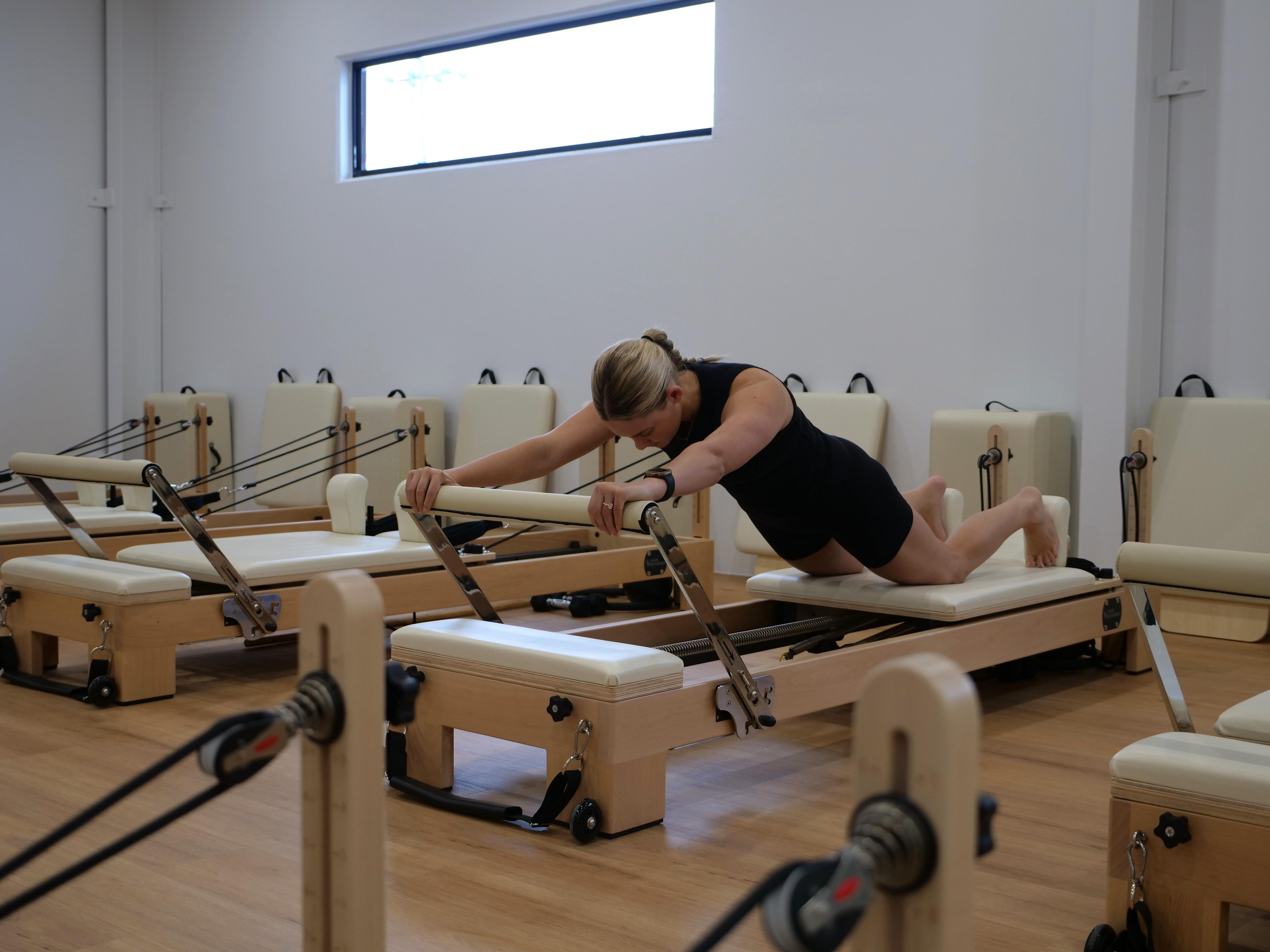 Young woman in black activewear horizontal on a cream and wooden pilates reformer equipment. 