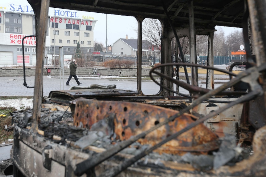 A man walks past a burnt out passenger vehicl.