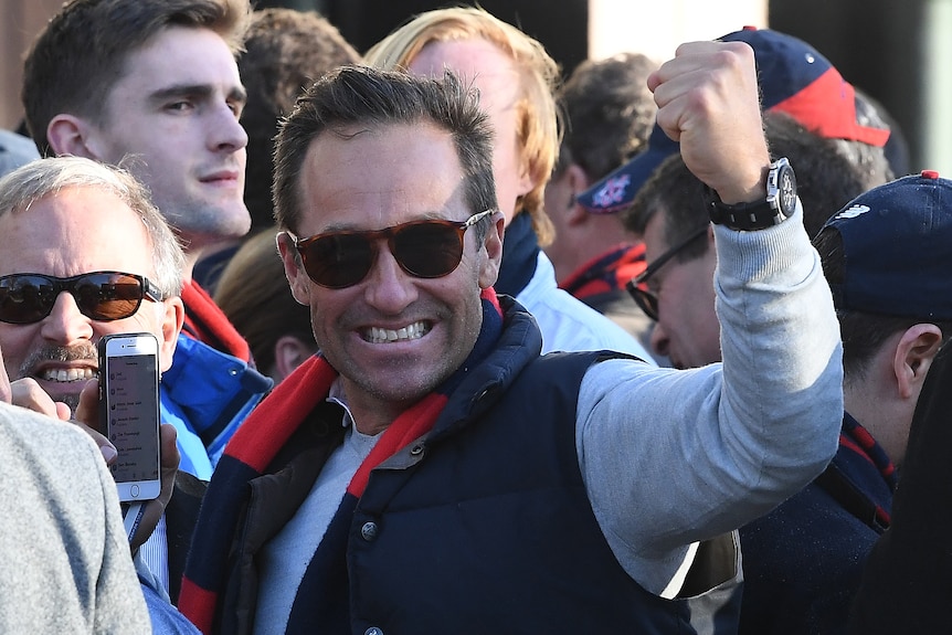 A man wearing a Melbourne Demons scarf holds a clenched fist in the air amid a crowd.