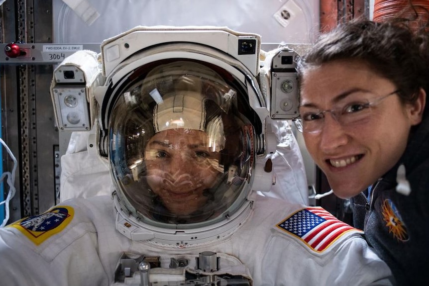 Two female astronauts smile for a photo one is dressed in a spacesuit.