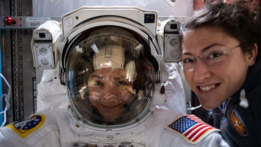 Two female astronauts smile for a photo one is dressed in a spacesuit.