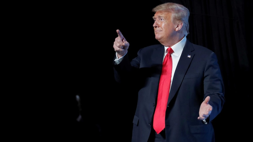 Blonde-haired man in black suit points with right hand