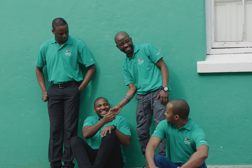 Four smiling Zimbabwean men stand and sit against a green shaking hands and laughing.