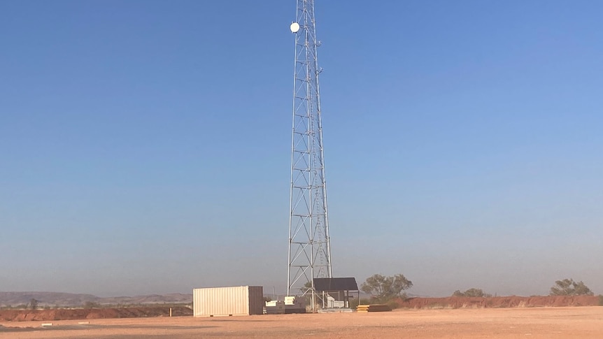 A large phone tower stands on dusty earth before a blue sky