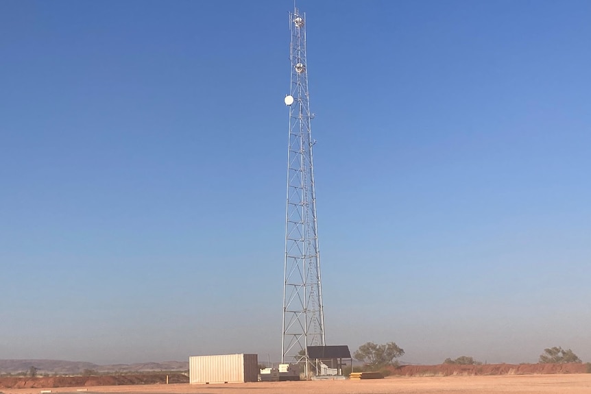 A large phone tower stands on dusty earth before a blue sky