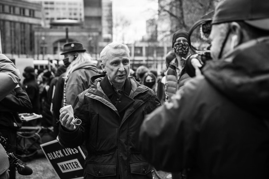 Black and white photo of man reporting to camera in a crowd of protesters.