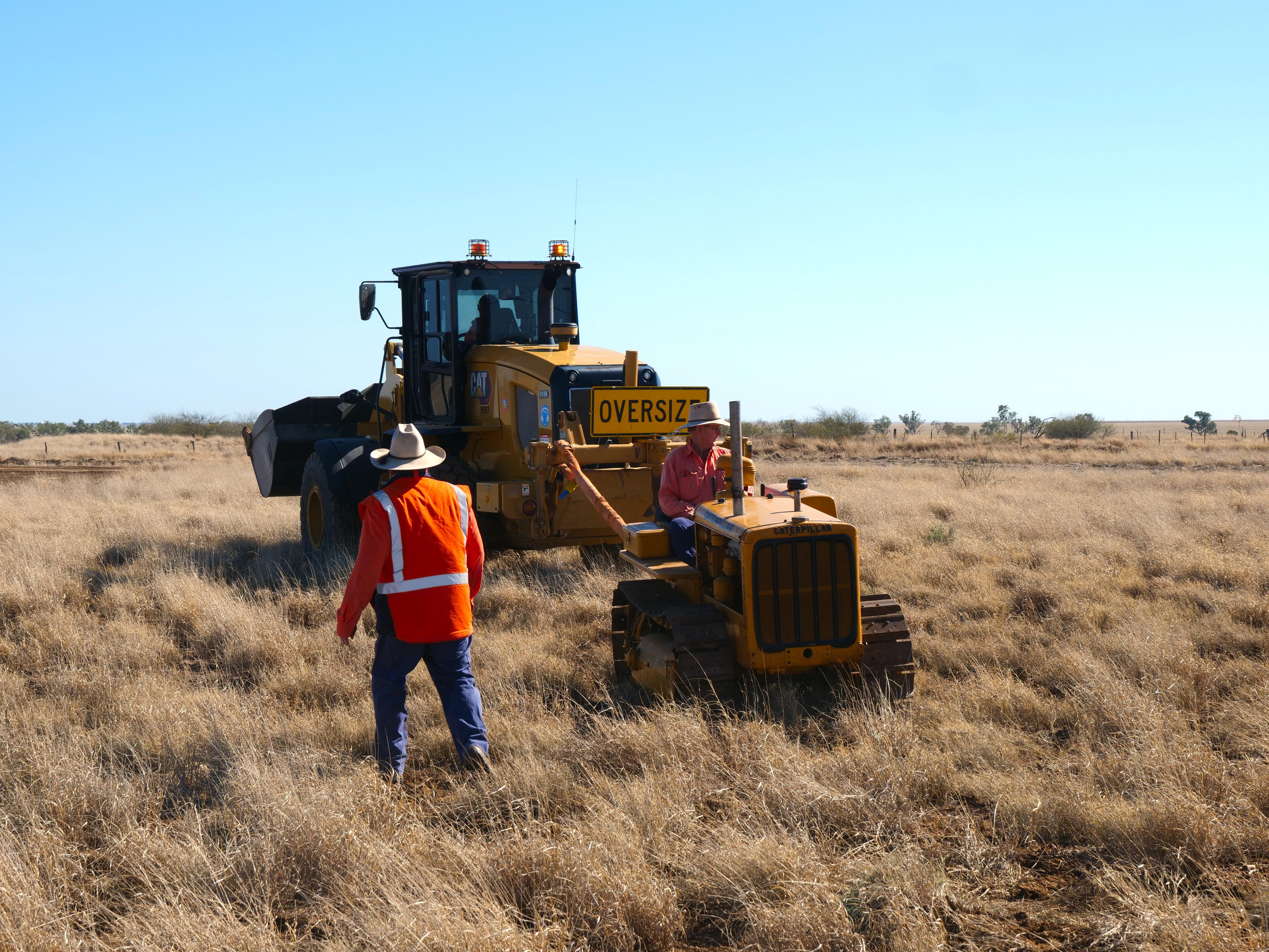 a man rides on a small tractor backwards through long grass, pulled by a larger tractor