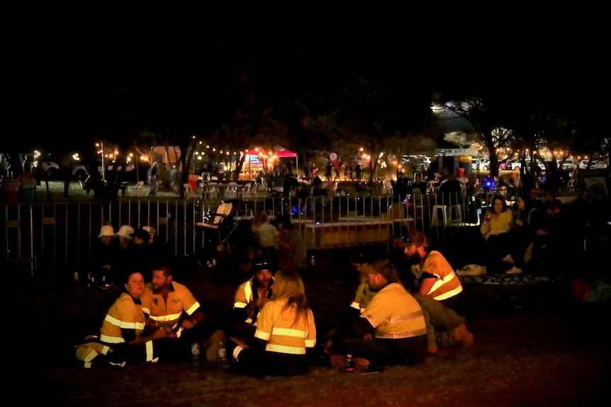 A group of people wearing high viz sit near a fair at night time.