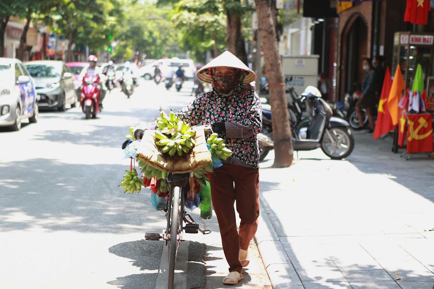 A woman wearing a face mask under a broad hat pushes a bike loaded with fruit along a road.