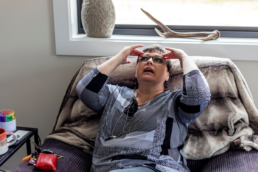 A woman with short brown hair and glasses wearing a grey patterned shirt sits of a couch with her hands rubbing her temples.