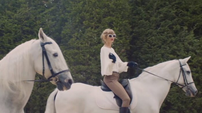 Singer Taylor Swift rides a white horse past large trees.