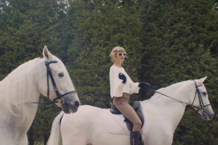Singer Taylor Swift rides a white horse past large trees.
