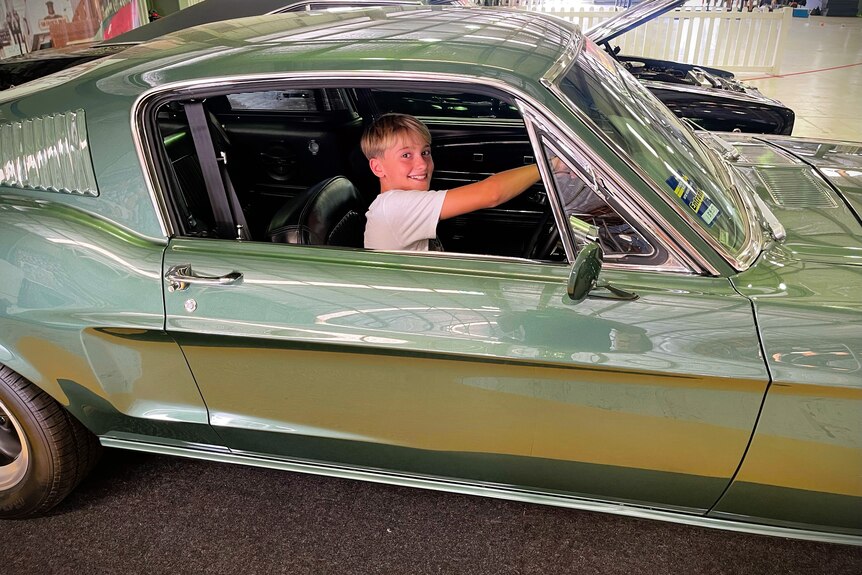 A boy with light, blonde hair smiling in the driver's sit of a green Ford Mustang car.