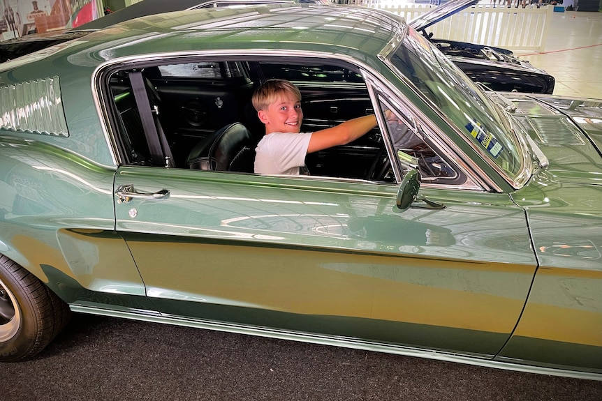 A boy with light, blonde hair smiling in the driver's sit of a green Ford Mustang car.