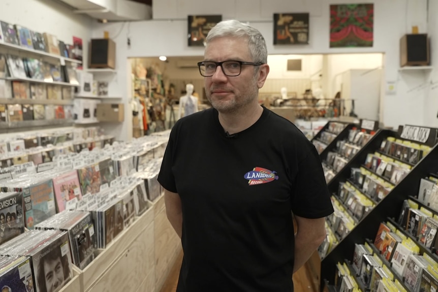 A man wearing glasses and a t-shirt stands in the aisle of a record store