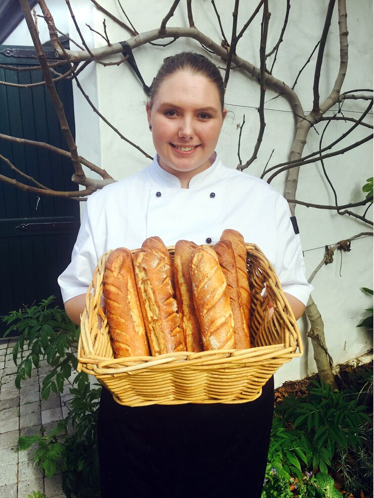 Young chef holding a loaf of bread outside a door