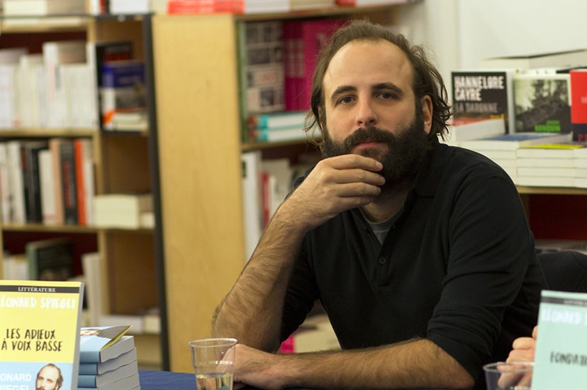 A man with dark hair, beard and hand held up to chin sits at desk with small stack of books inside bookstore.