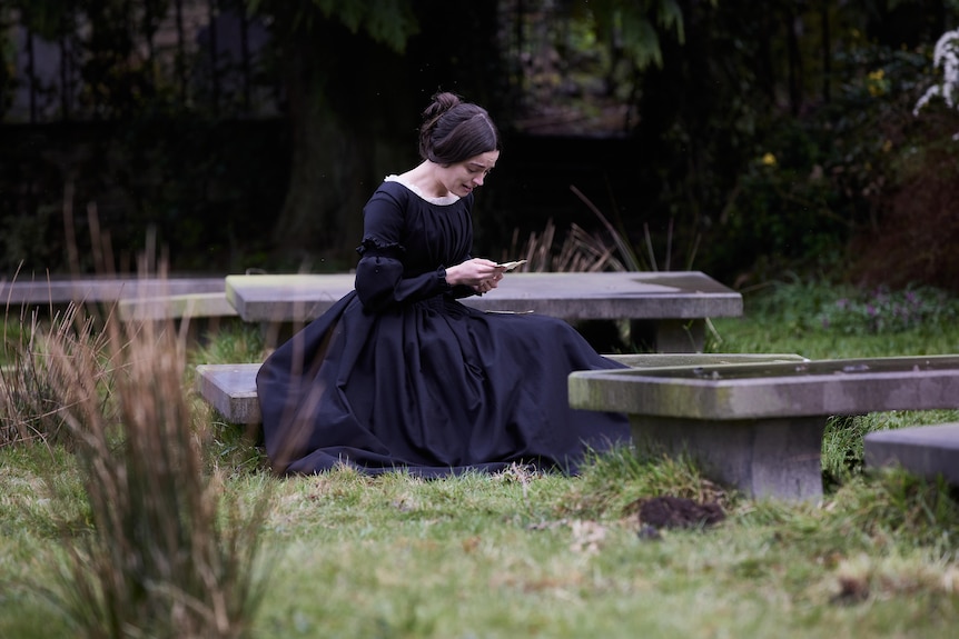 White woman with dark hair pulled back wears an 1800s style black dress and sits in a garden reading a letter while weeping.